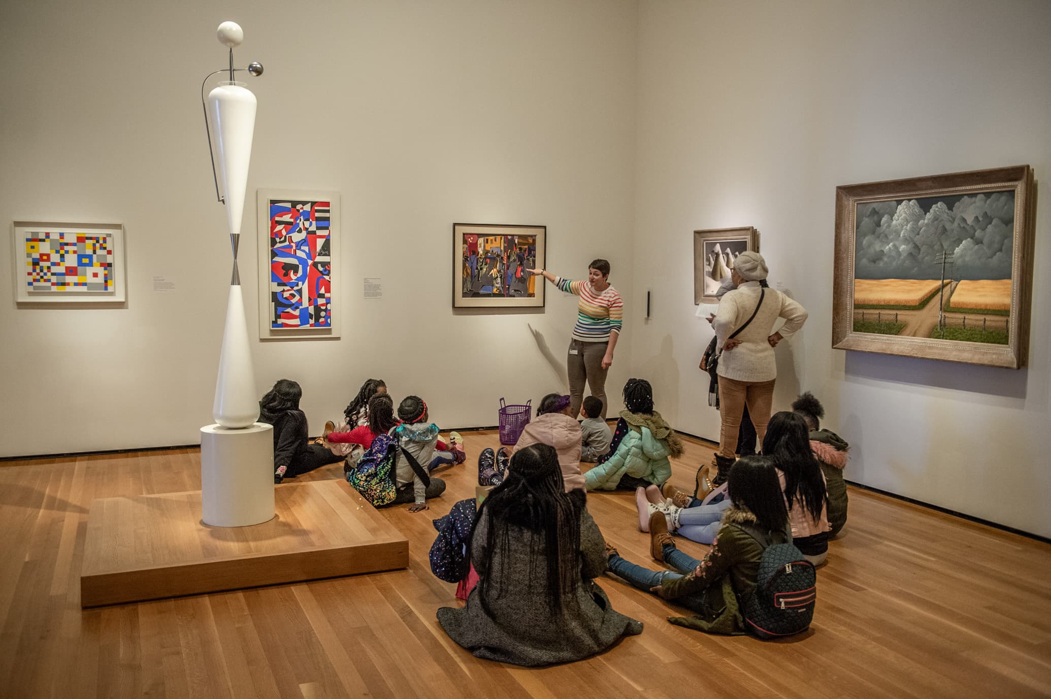 tour guide describes painting to group of children & adults, seated on the floor of a museum gallery