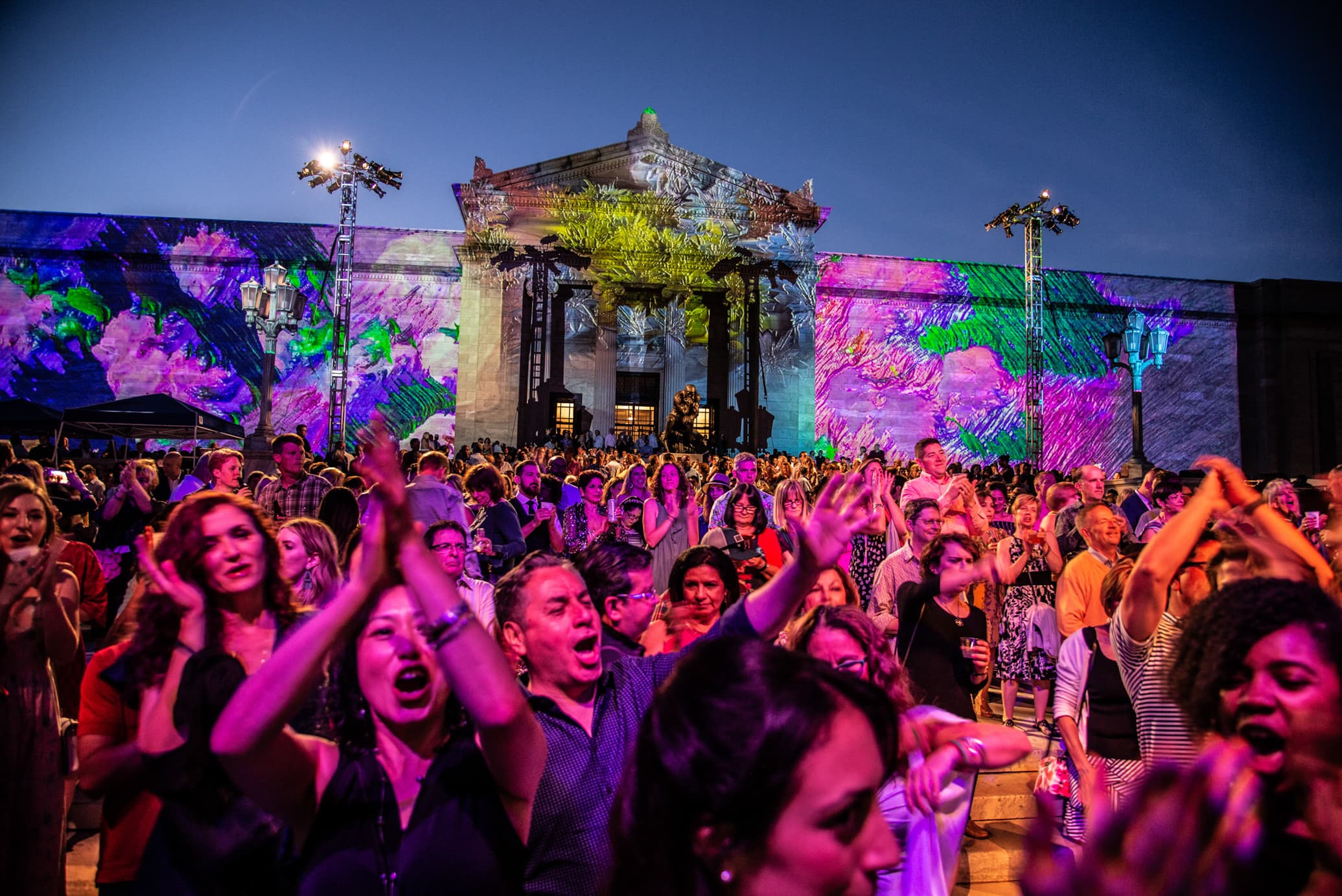 large dancing crowd in front of museum facade, lit by colorful projections