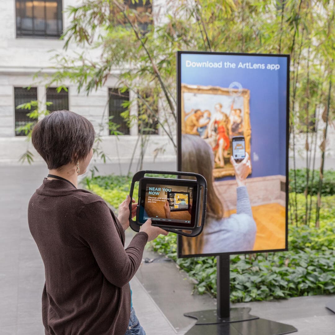 A person standing in front of an ArtLens app sign while using the app on an ipad.