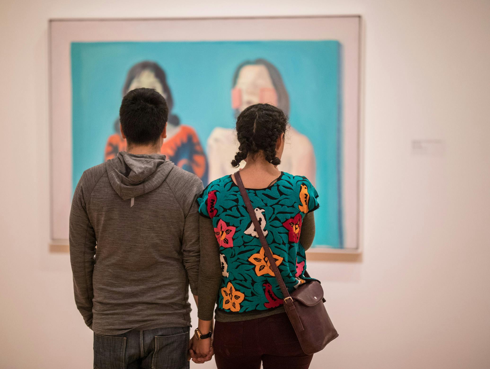 A back of a man and a woman holding hands while looking at a painting