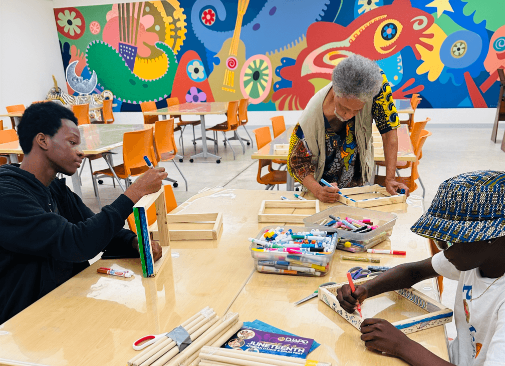 A teacher with two students decorating African box drums at a studio table.