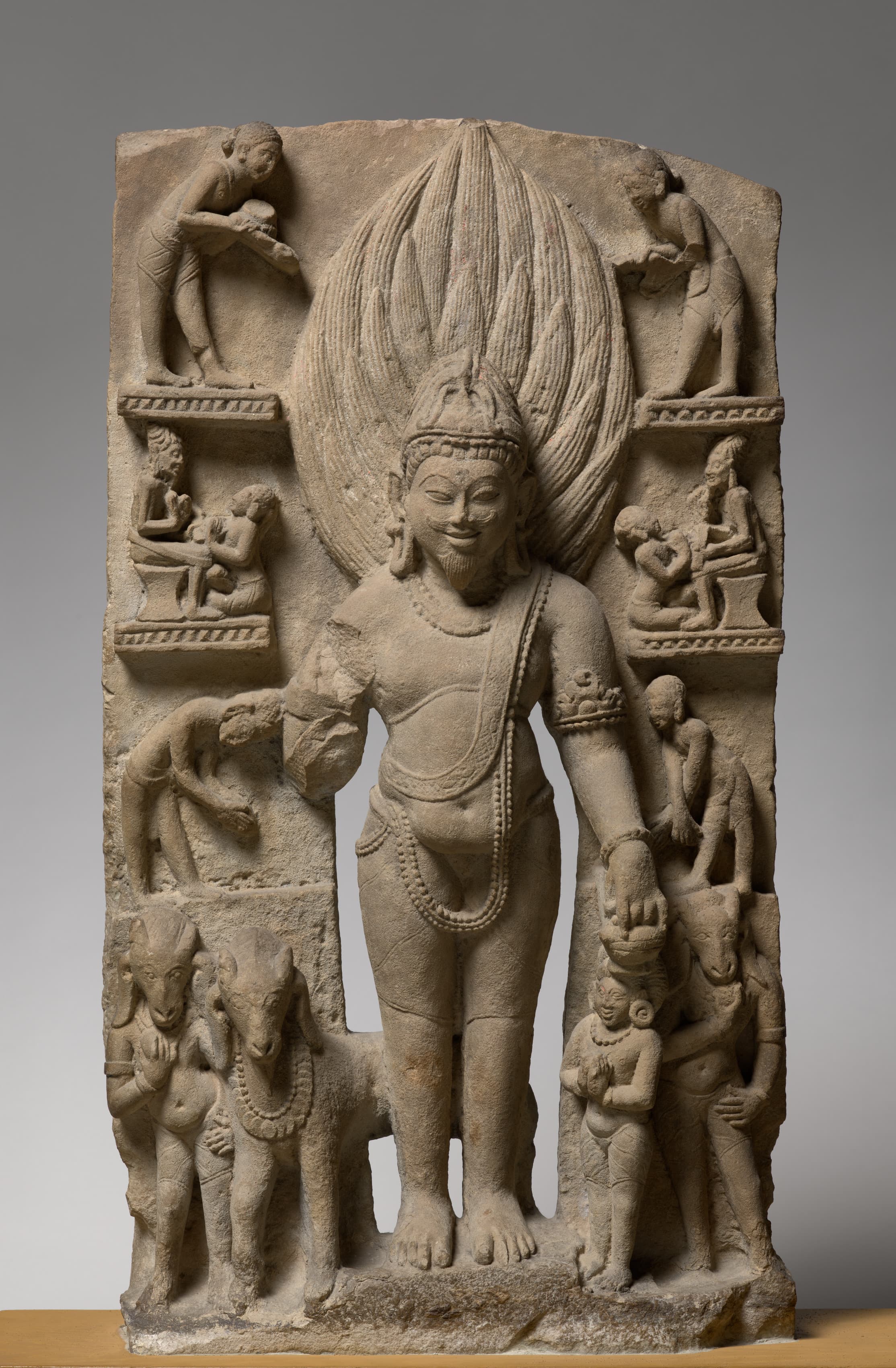 A gray-brown sandstone full-body sculpture depicts the god Agni with flames flaring behind his head and flanked on either side in four rows by figures a fifth of his size, then a third of his size at the base, including a goat and figures with human bodies and goat heads. He wears a crown and jewelry, has a mustache and small pointed beard, with his left arm hanging down and the other broken off at the elbow.