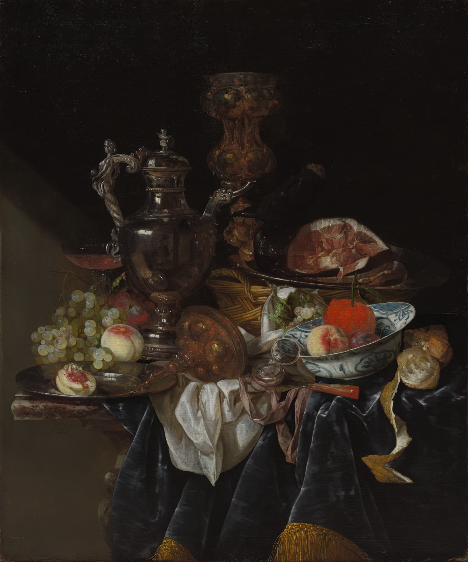 A vertically oriented oil painting depicts a still life on a table draped in dark blue velvet and white linen. A tall silver ewer and amber glass vessel stand behind a blue and white bowl of peaches. To our right, a platter holds sliced ham and bread, while a curled lemon peel hangs over the edge. Green grapes cluster on our left. Strong light creates brilliant reflections and deep shadows across the composition.