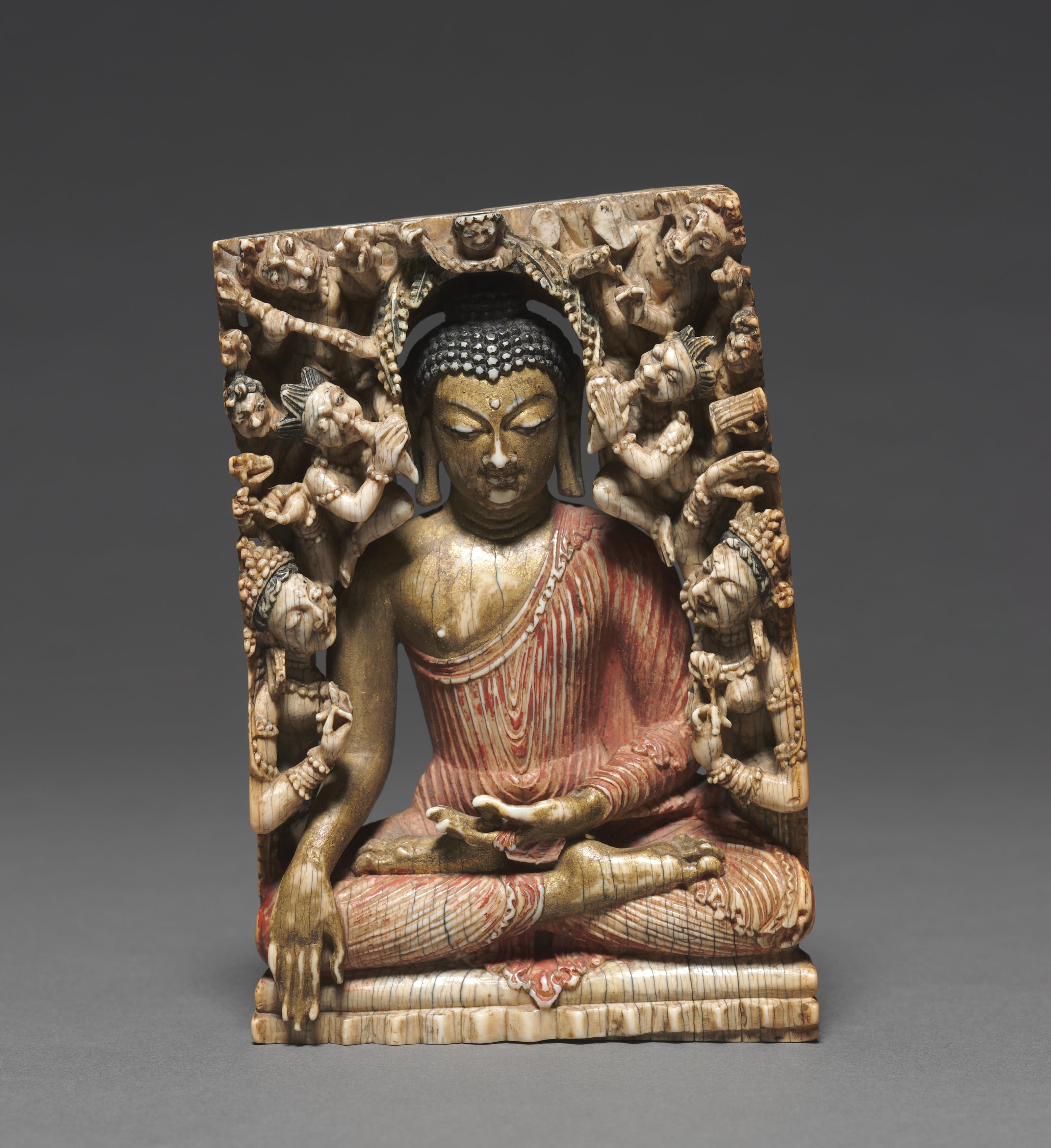 A painted, light brown ivory sculpture depicts the Buddha seated cross-legged while women and demons a fifth his size arch over him, the edges cut into a sharp, slightly curved rectangular sculpture. The Buddha places his left hand face-up on his lap, his right draped over his right knee, and wears a red, rippling robe over his left shoulder. His skin is painted gold and his black hair features rows of knobs.