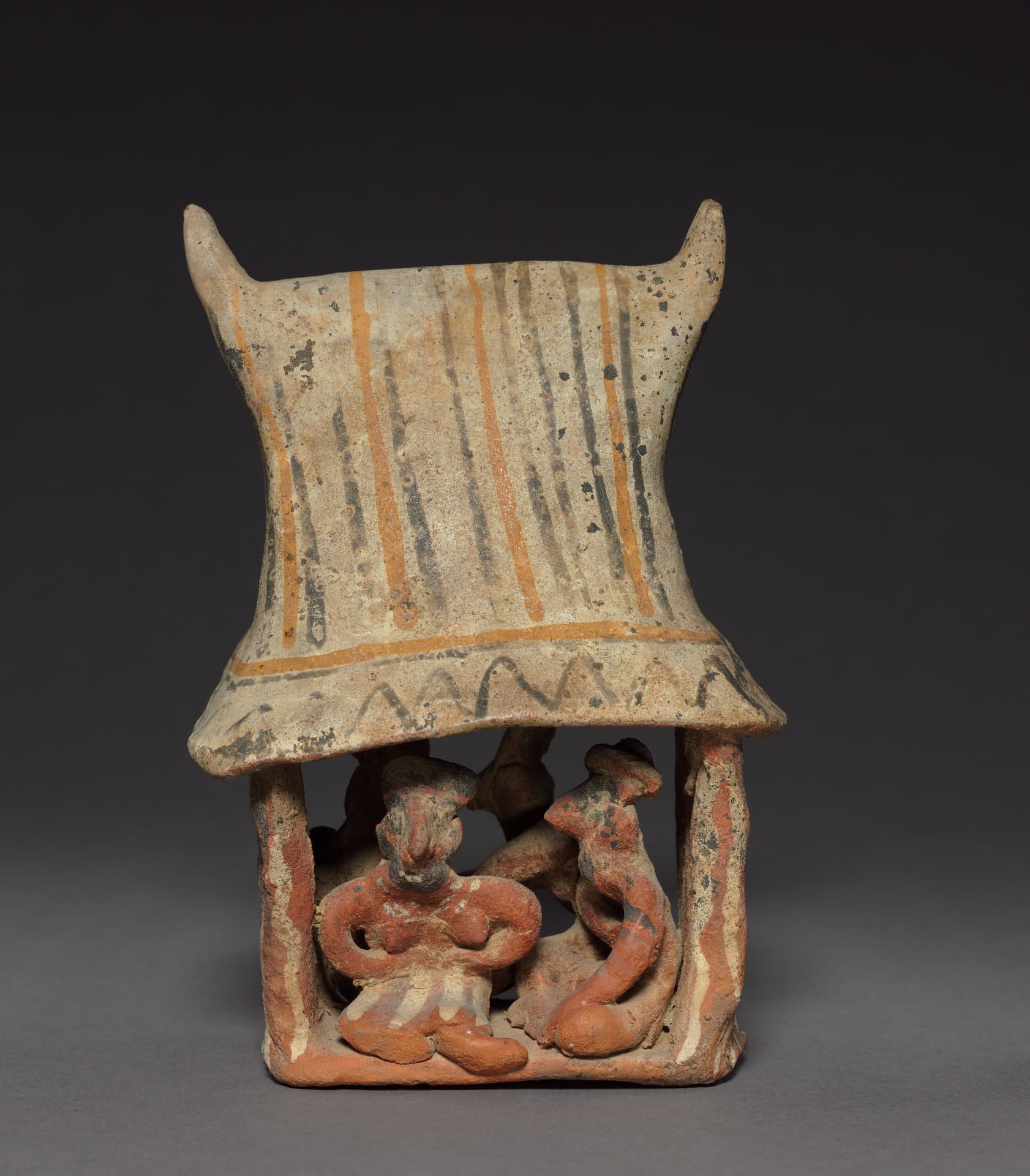 An earthenware sculpture in earthy creams, oranges, and reds depicts an architectural scene. A tall, saddle-shaped roof with upward-curving points features vertical stripes and a black zigzag border. Below, two thick pillars support the structure, housing reddish-brown figures. A central figure sits facing forward with arms folded across their chest, while another sits in profile nearby. The figures are nestled within the open base, framed by the dominant, decorated canopy.