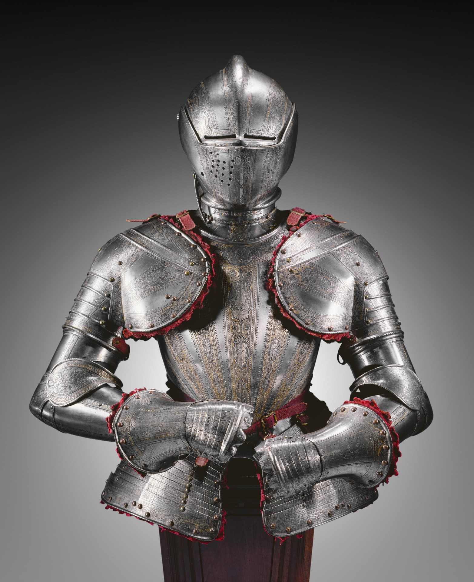 A half of a set of steel armor including the helmet, shoulder, arm, chest, and torso pieces is detailed with etchings of patterns and figures. It also contains a reddish-purple belt and red velvet fittings visible at the edges of the armor.