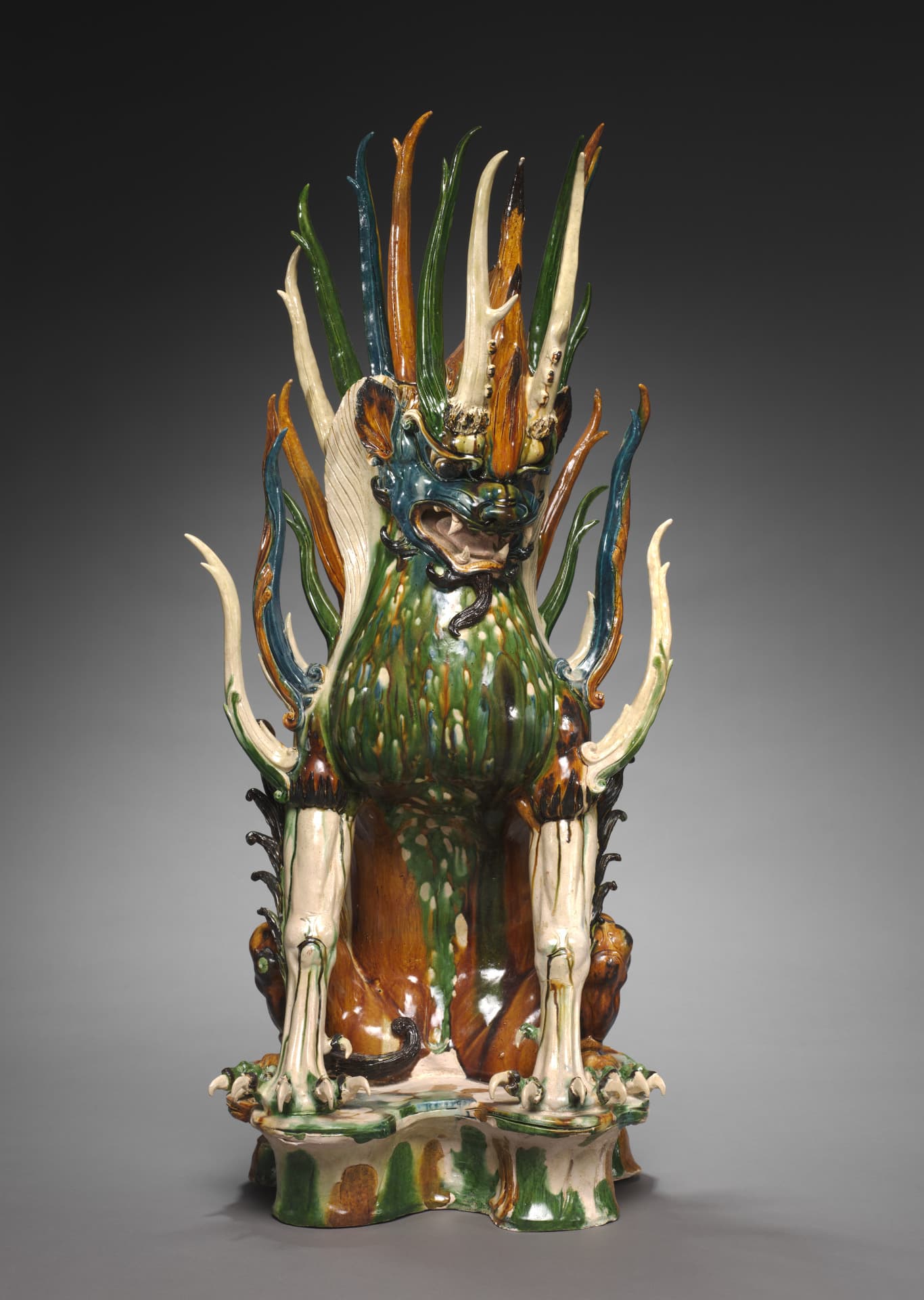 An earthenware sculpture, glazed in green, orange, blue, and cream, depicts a mythical beast with a blue animal face and claws, seated on its hind legs. Its mouth is open, baring its fangs. Antlers spike upward from its head and further spikes from its shoulders and the ridge of its back.