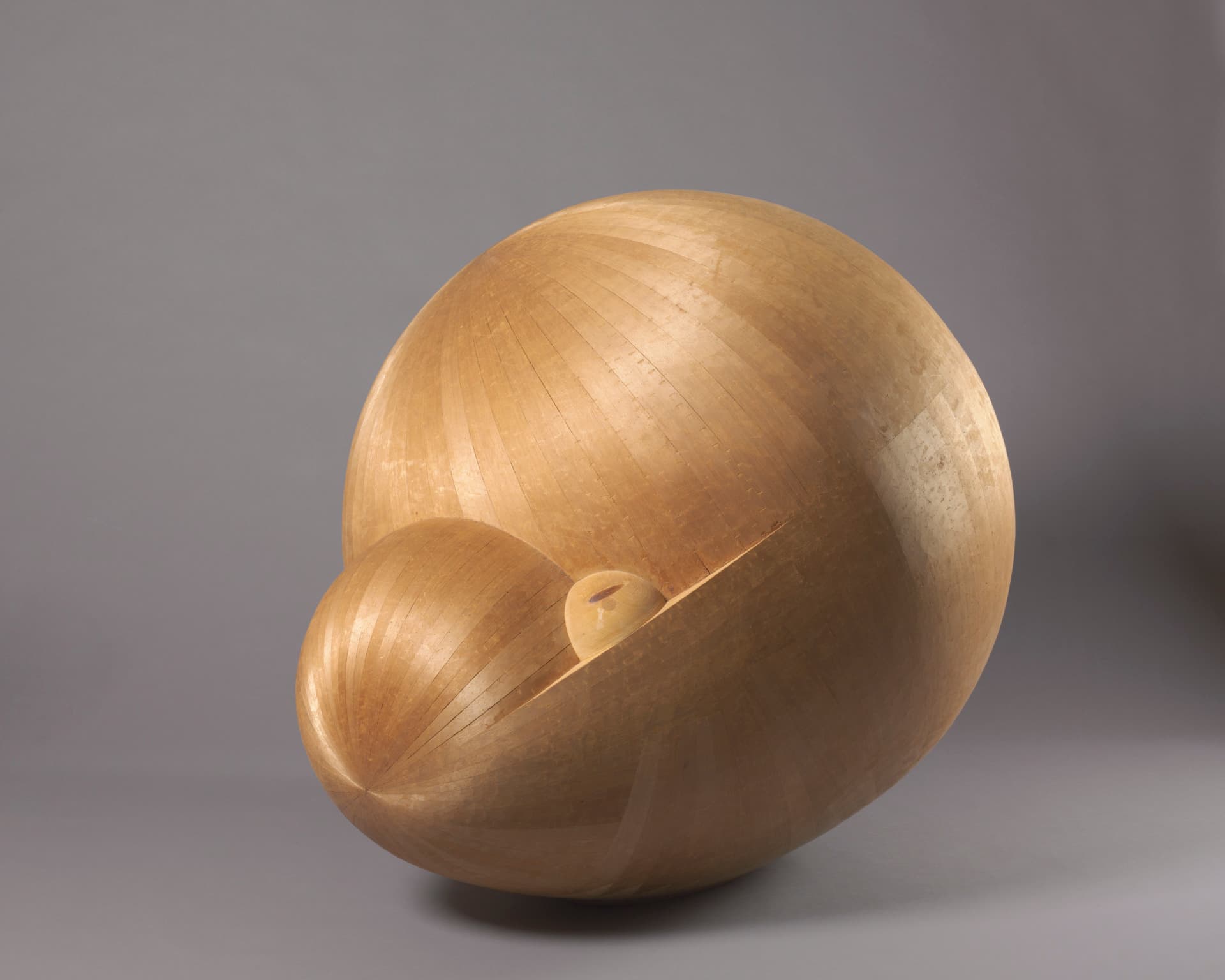 Three blond wood spheres, made up of thin smooth panels, join together. At the largest sphere's low edge, almost touching the ground, half of another smaller sphere is attached. Nestled between the two is a very small third sphere.