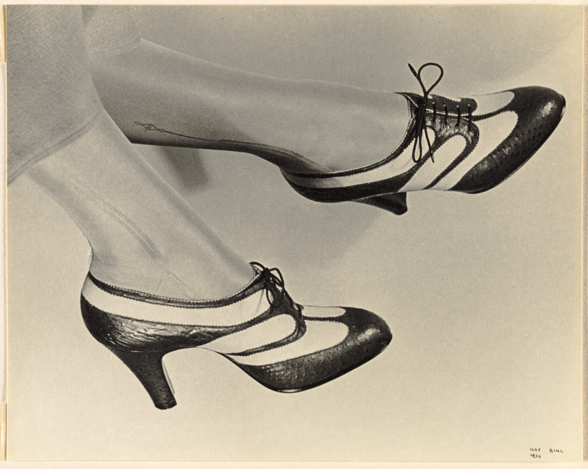 A horizontally oriented black-and-white photograph shows two feet wearing stockings and heels dangling in from the upper left corner. Framed against a neutral, light background, the heels mix leathery black portions with smooth, white, elongated blobs and have fine laces at the top. The left foot kicks out horizontally, tilted in slightly where the laces can be seen more clearly. The right foot hangs down at a diagonal, highlighting the profile of the heel.