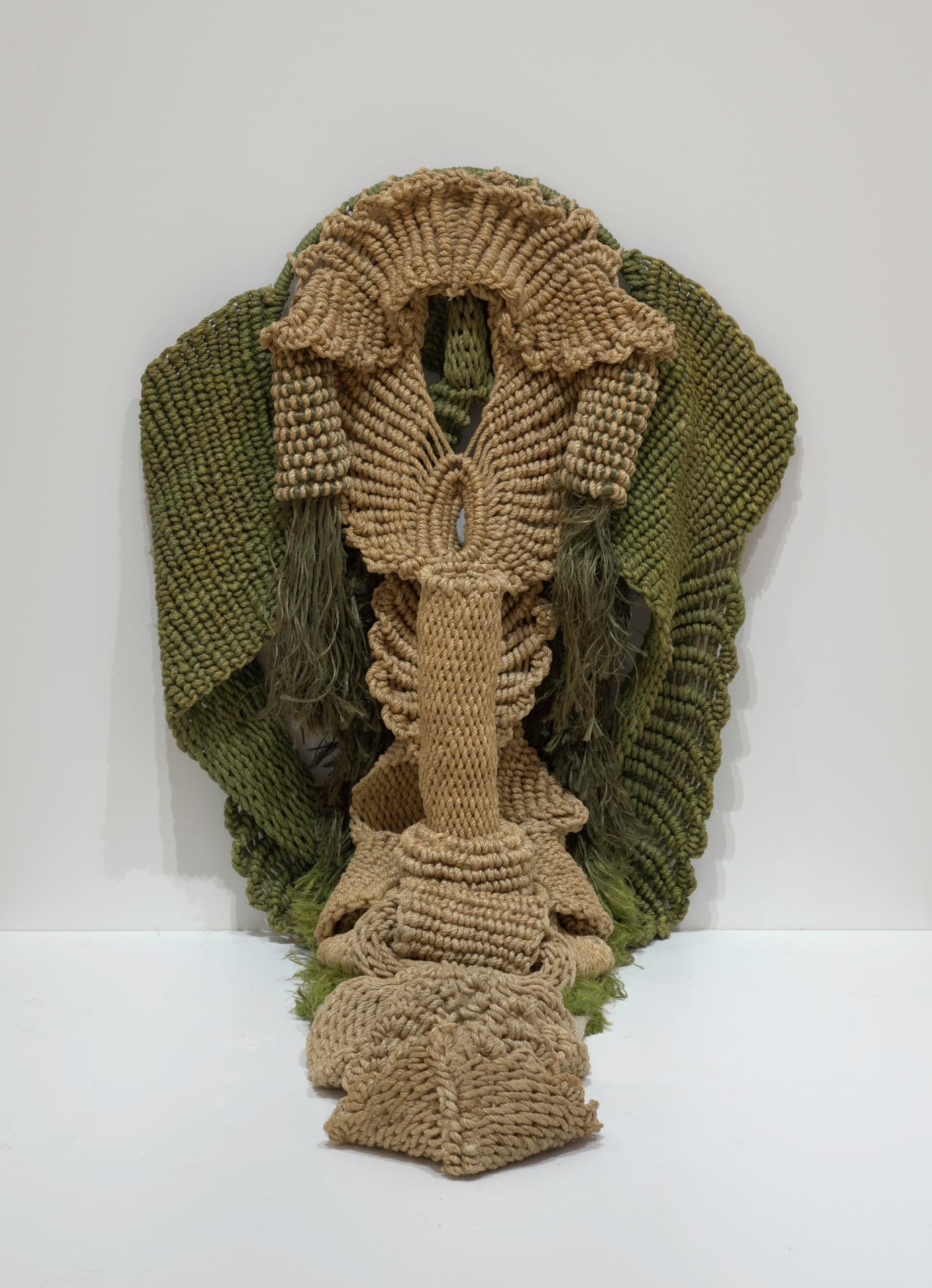 A knotted hemp rope in green colors has rippling, bumpy edges like a woven rug and is reminiscent of a cobra hood. A brown-green strip extends down and piles on the floor before lifting up into an elongated neck from which riblike protrusions stick out. Dark green knotted hemp rope flares on either side from behind the cobra hood.