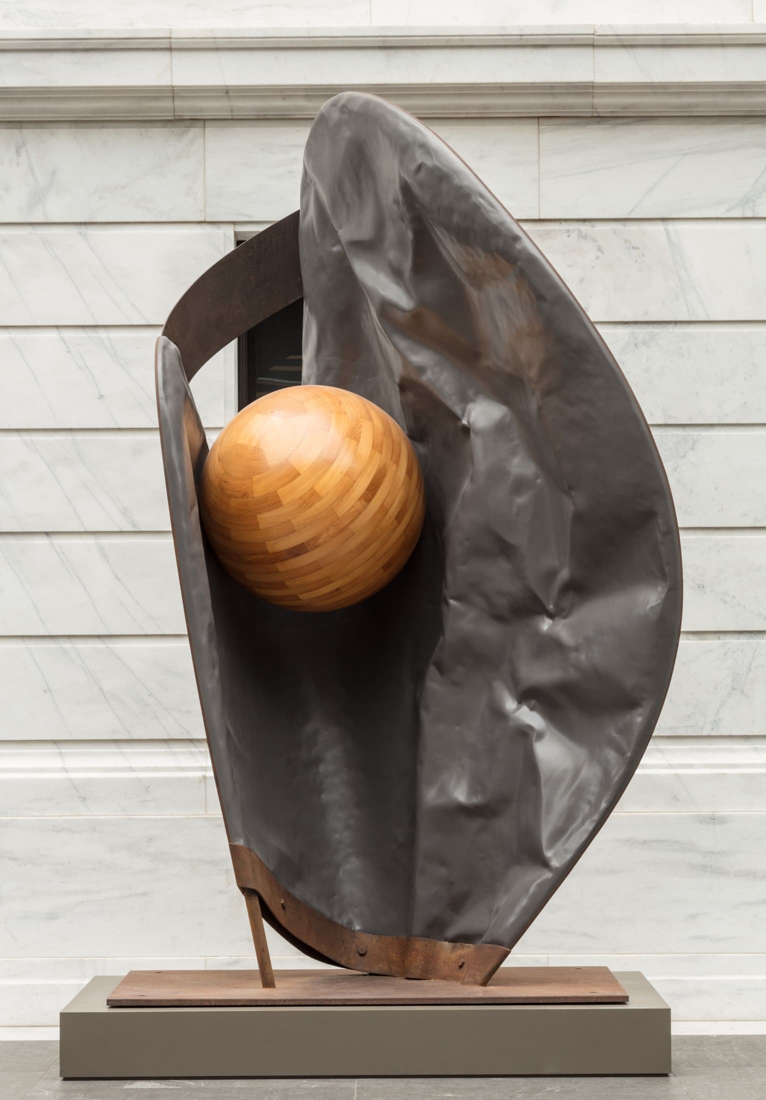A lead and steel gray-brown baseball mitt is tilted up on a base and holds a wood ball made of curved planks of light brown, striated wood. The glove warps around the ball, a rectangular band connecting the body of the glove to the thumb.