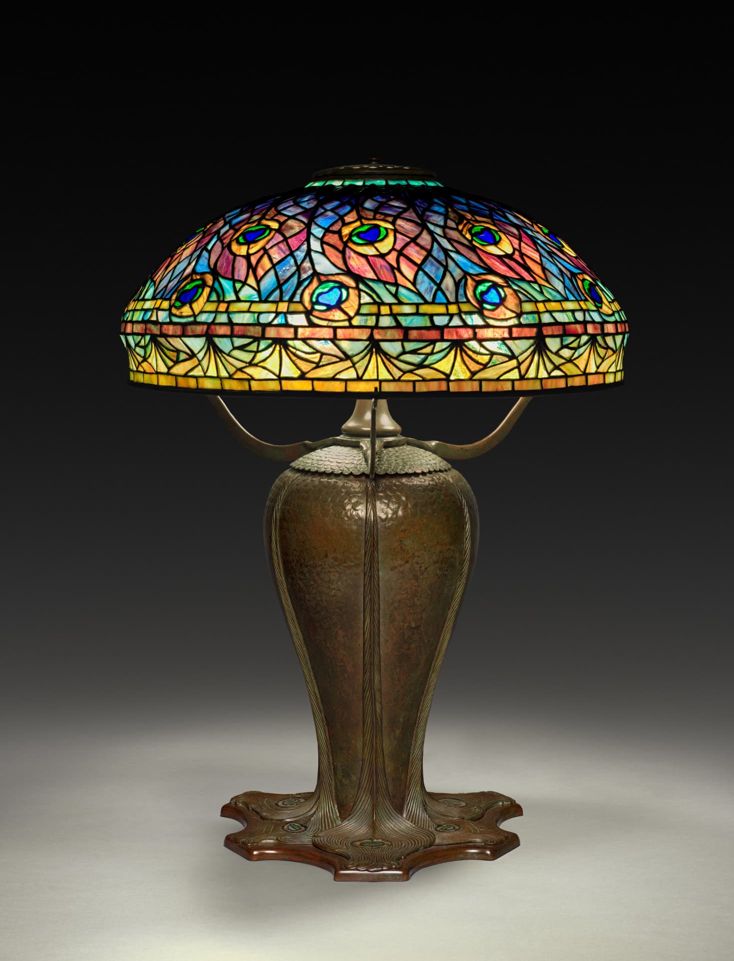 A table lamp features a colorful, glass lampshade resembling peacock feathers over a bronze body reminiscent of a peacock's body, having a broad shoulder with scalloped feather patterns, narrowing before flaring at the base with larger peacock feathers. The lampshade, a shallow dome over the lamp, is patterned with blue, orange, red, and green glass. Winding, dark metal lines connect the various pieces of glass together, light from the bulb within highlighting the glass' striated shades.
