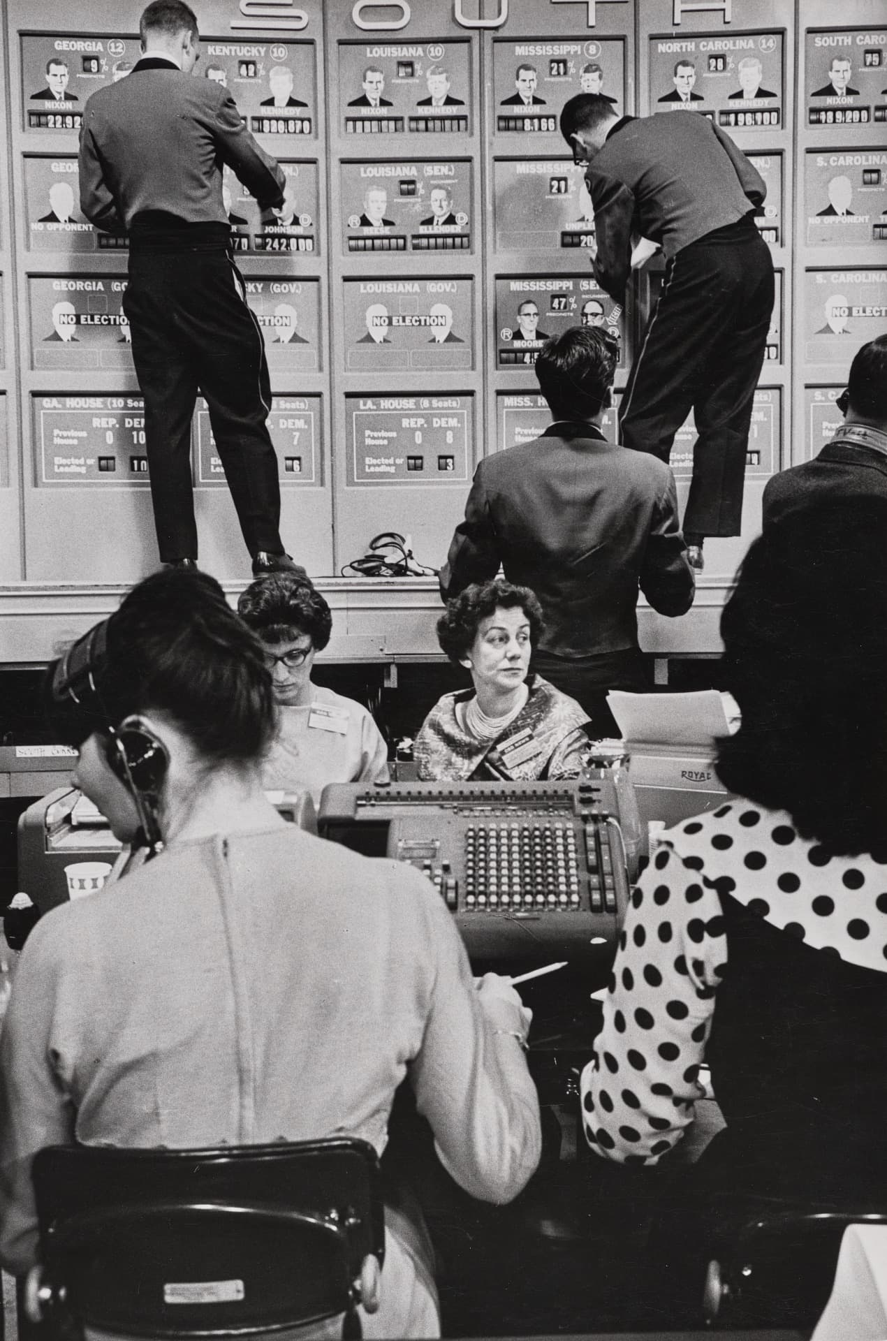 A vertically oriented black-and-white photograph depicts a crowded newsroom. In the foreground, two women sit at desks with their backs to us, the woman on our right wearing a polka-dot blouse. Behind them, another woman sits looking toward our right. In the background, two men on a platform update large wall charts featuring portraits of candidates Nixon and Kennedy, state names, and numerical election results beneath the word "SOUTH."