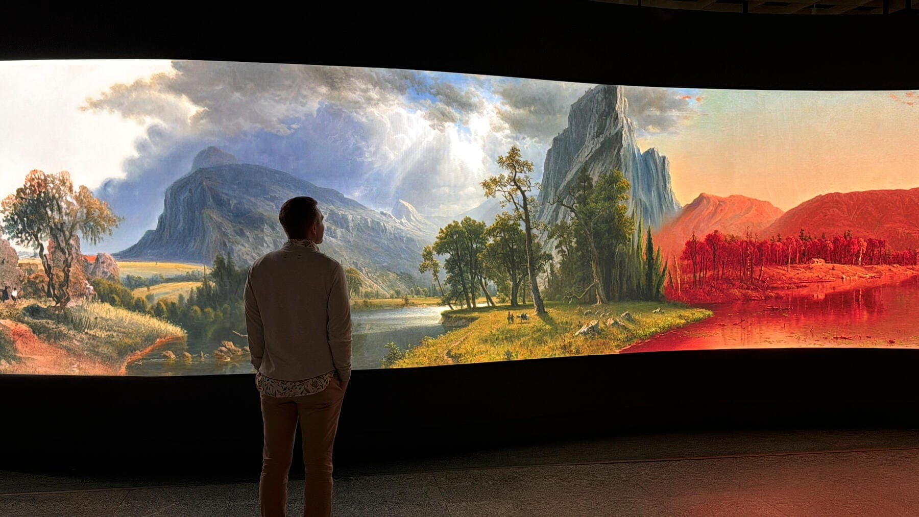 a man standing in front of large panoramic LED screen displaying painted river and mountains