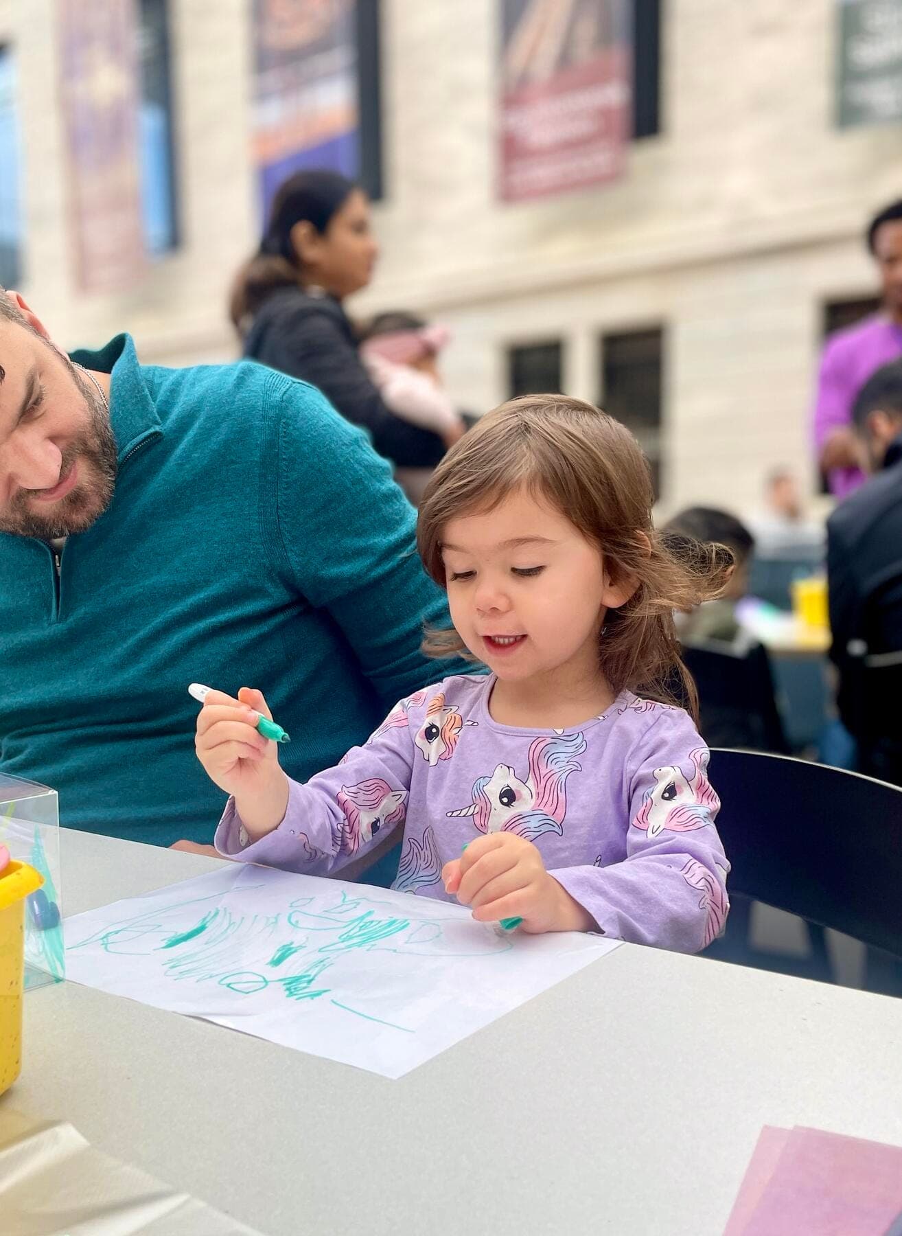An adult and a child sitting together, the child is drawing on paper using a marker.