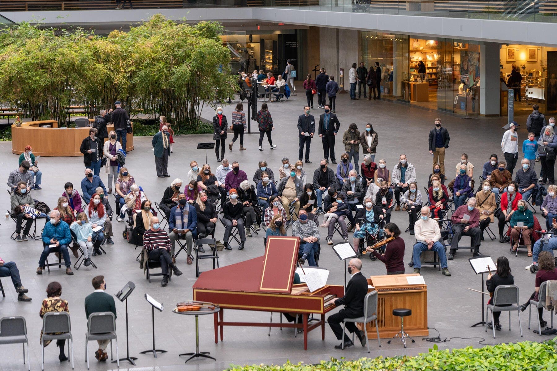 A large gathering of people in the museum's atrium sit around a harpsichord and small organ, listening to a live performance.