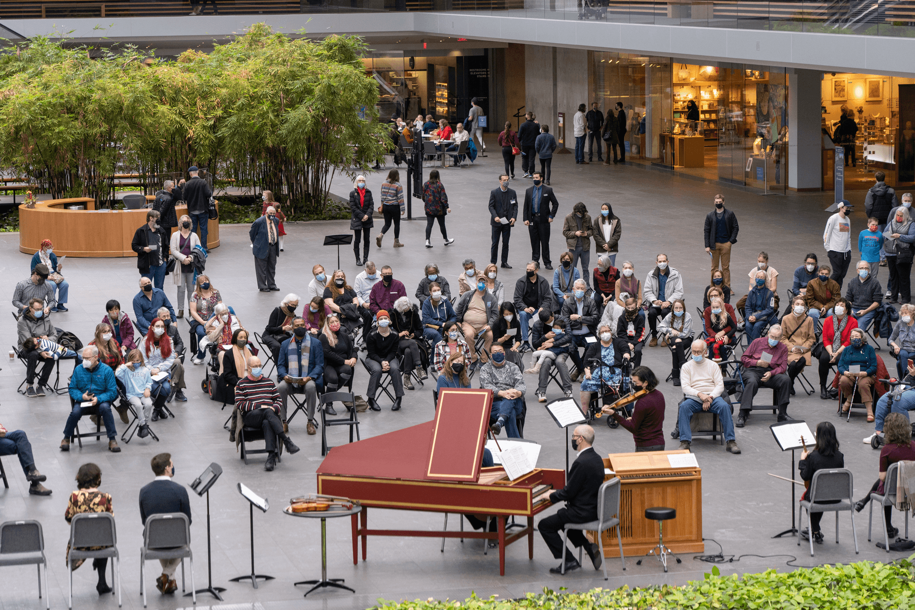 A large gathering of people in the museum's atrium sit around a harpsichord and small organ, listening to a live performance.