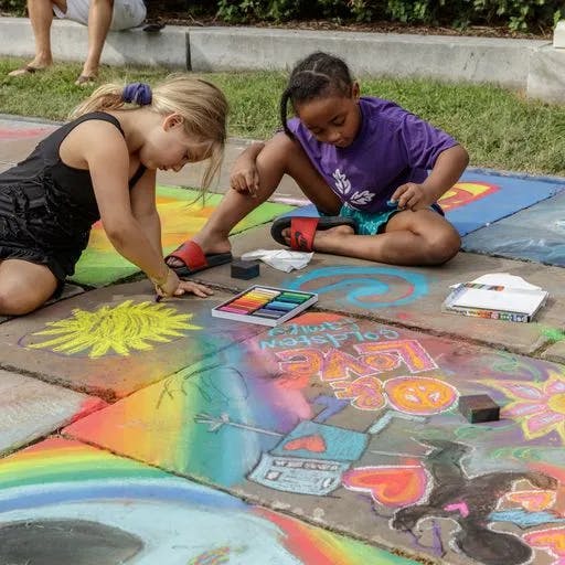 Two young girls drawing with colorful chalk on the side walk. There are already chalk drawings on the ground of a robot and the word "love"
