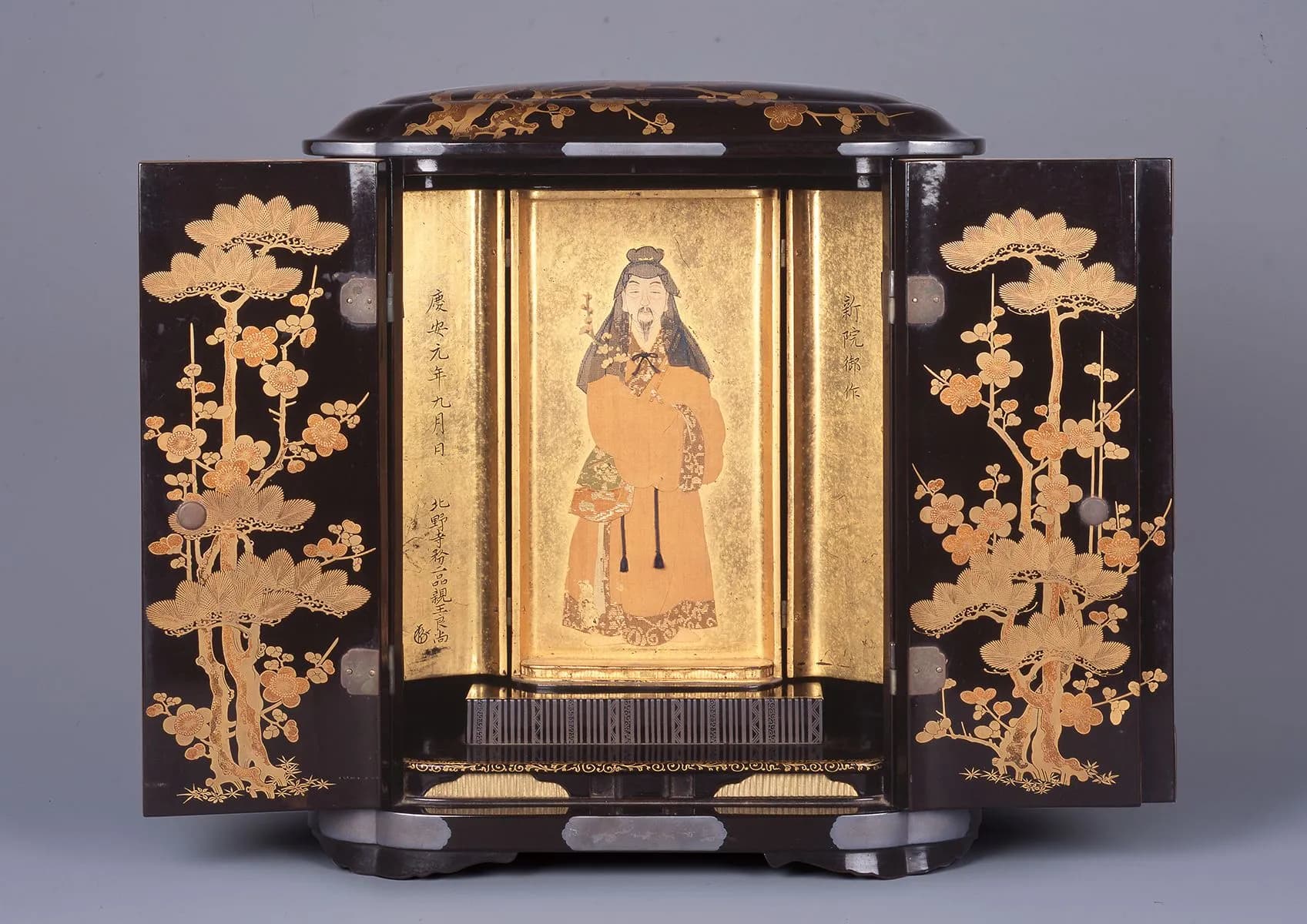 Devotional shrine with doors open to reveal the image of a figure facing us in pale orange robes and against a gold background. On the inside of the shine black doors climb gold plant designs with fine detailing.