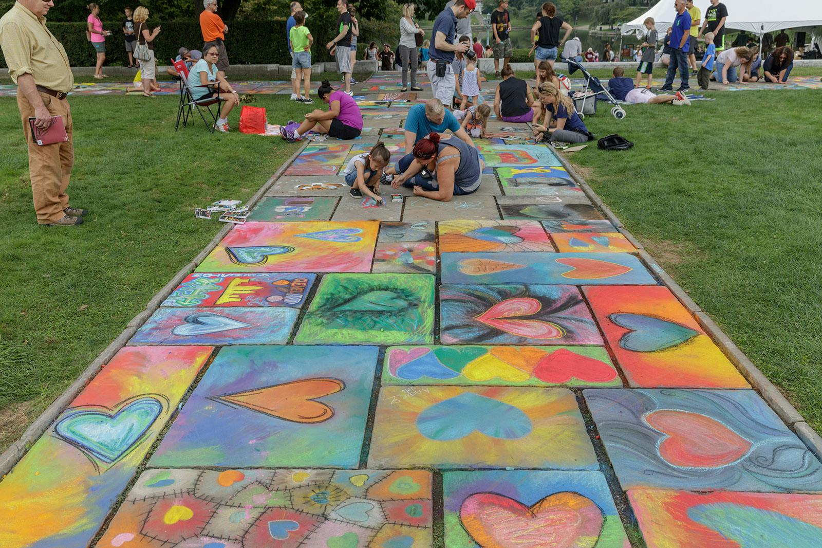 Colorful chalk drawings on stone walkway; children and adults draw on stones in background