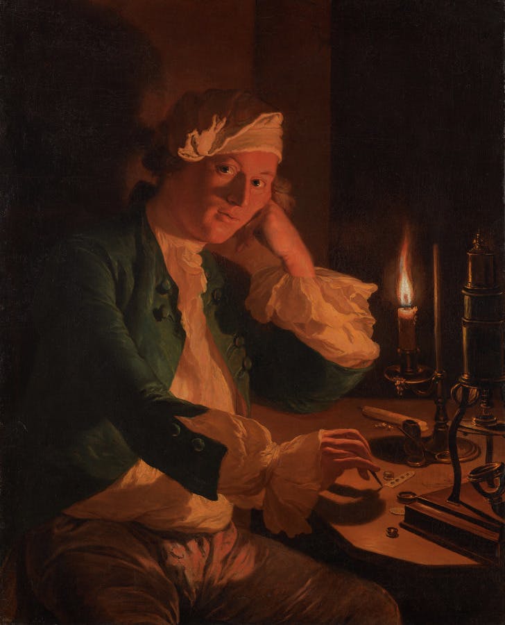 A man lit by a single candle wearing a cap sits at desk, His face rests on his hand, the direction of his gaze appears to be aimed back at the viewer.  