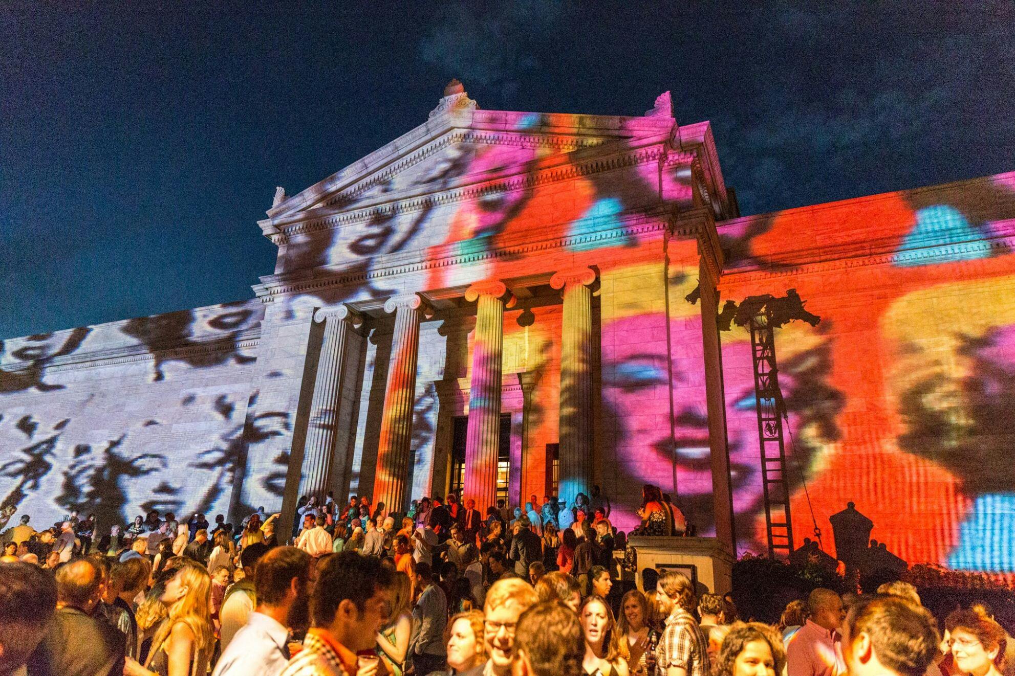 a crowd of people gathered outside the CMA in celebration, with art projected onto the building behind them