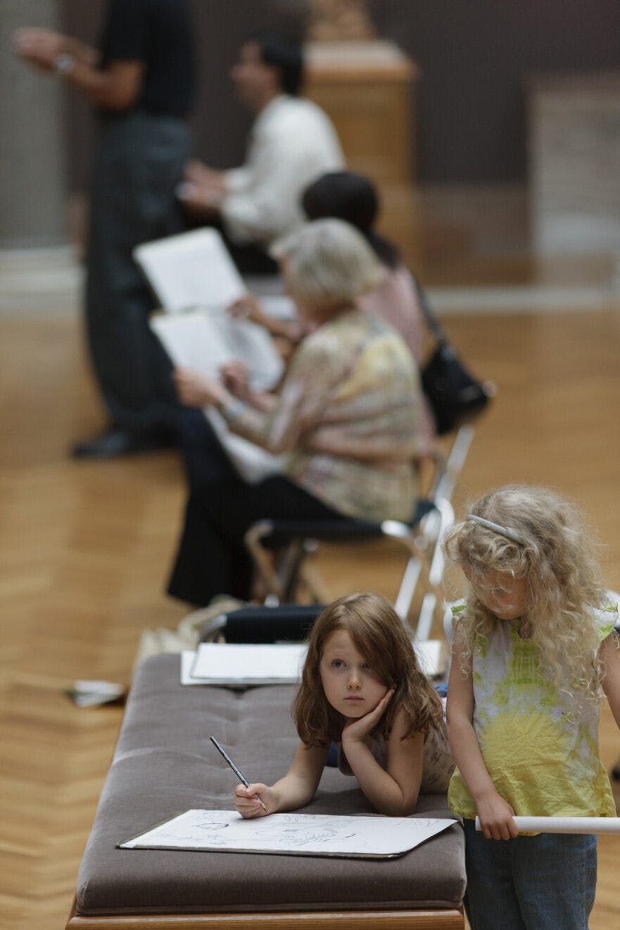 young children sitting on a bench at the CMA drawing on a large sheet of paper