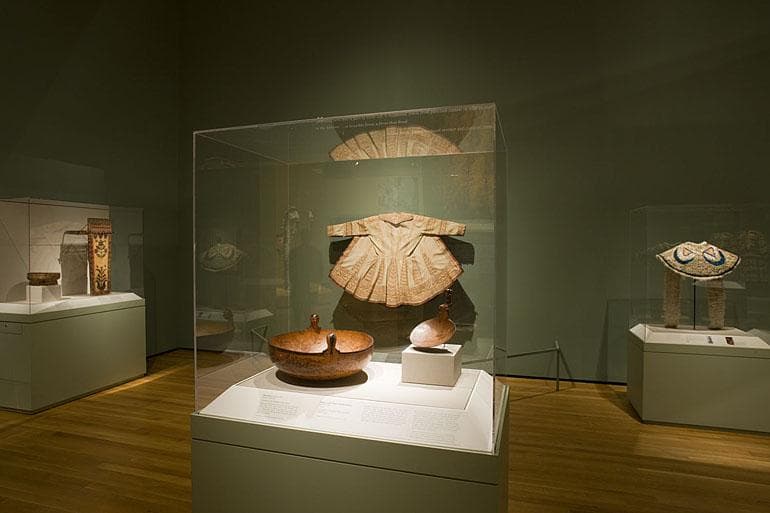 gallery view of a dramatically lit case with a vessel and a garment on the wall
