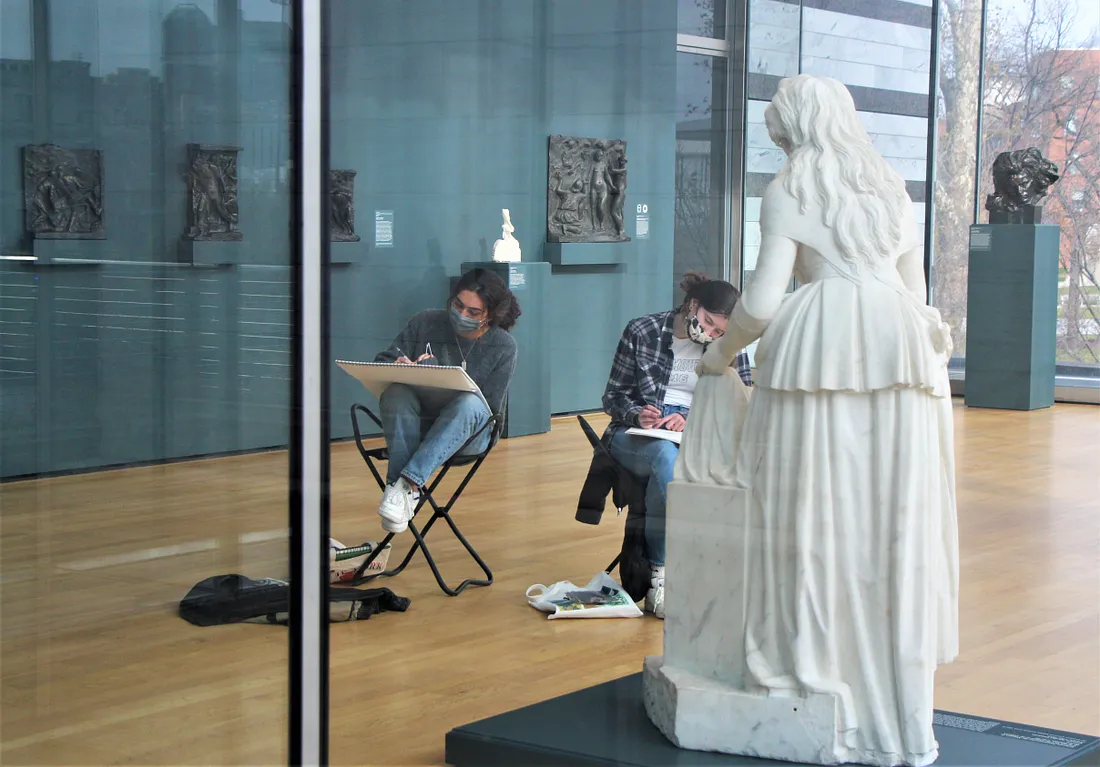 Visitors sketching in the galleries in front of a white statue of a woman with long hair