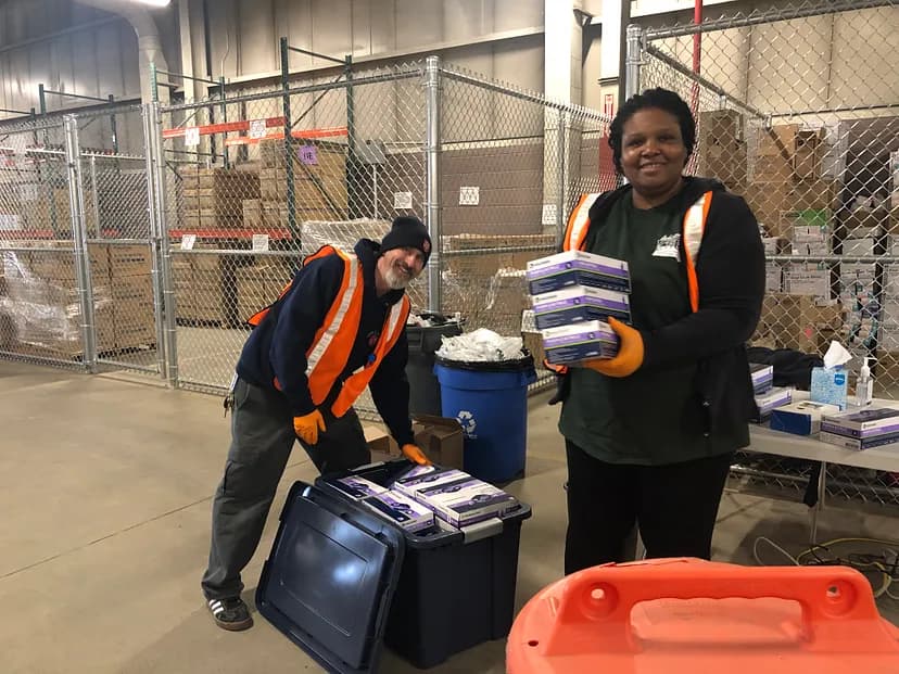Two people wearing orange vests in a warehouse and holding boxes.