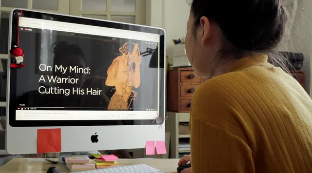 Image of a person looking at a computer screen with a video playing, the words "On My Mind: A Warrior Cutting His Hair" on the screen.