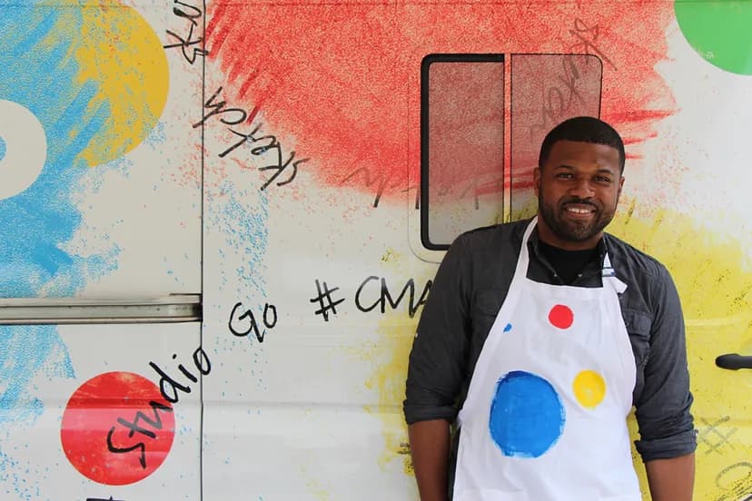 Person in apron in front of van decorated with colorful splotches, the words "Studio Go #CMA" visible.