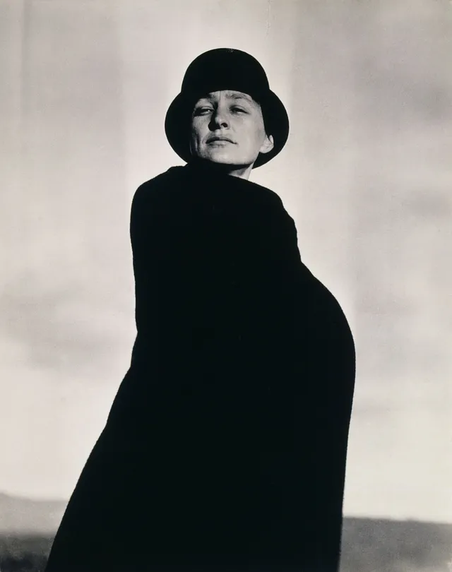 Black-and-white photograph of a woman in a black coat and hat, framed by a cloudy grey sky.