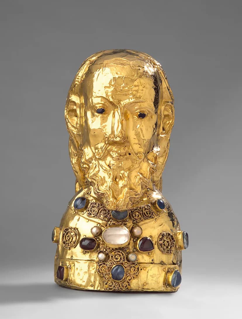 Wood sculpture coated in gold of the head of a bearded man with black stones for eyes and jewels around his neck and across his chest, a large white jewel at the center.