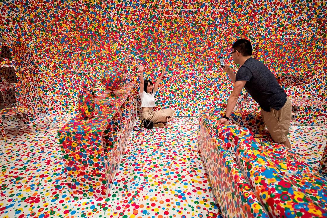 Room with wall, floor, shelves and couch all covered in green, blue, yellow, red, and orange dots in which one person takes a picture of another.