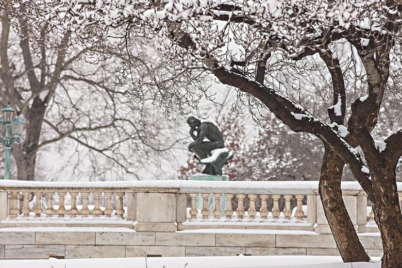 A bronze sculpture of a man resting his head on his chin, with snow on his back and seen through snowy trees.