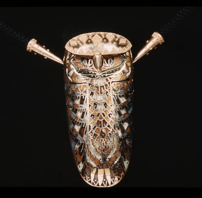 Oval-shaped gold pendant owl inset with enamel and blue, burnt orange, and yellow details and with two cones protruding from either side of the head.