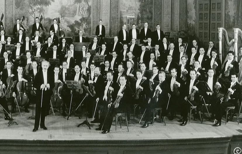 Musicians in an orchestra on a stage, wearing suits and holding their instruments as they look at the camera.