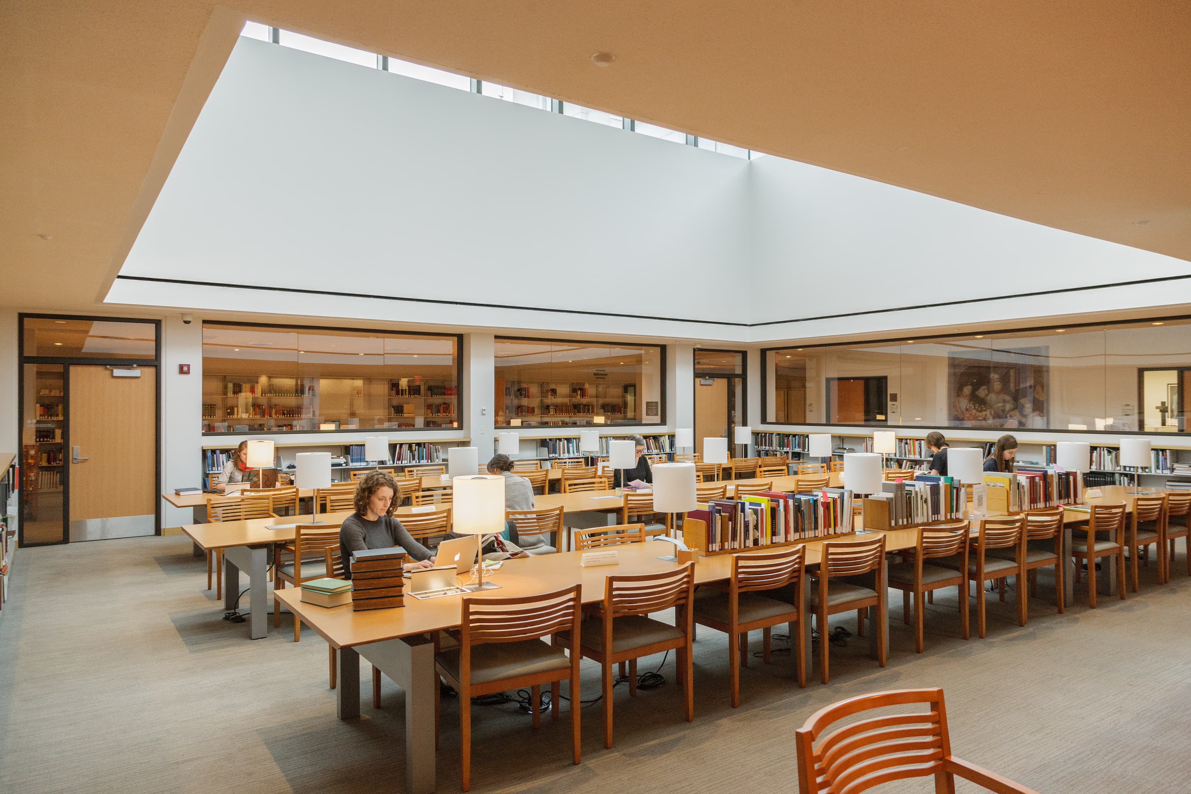 Light wood tables and chairs in a library with a few people sitting at the tables.