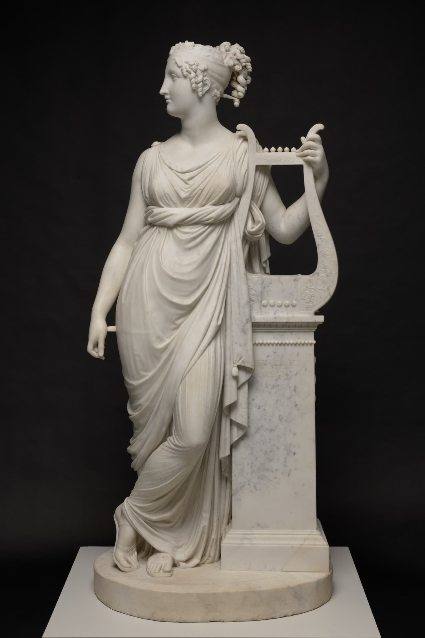 A white marble sculpture depicts Terpsichore Lyran, a muse standing next to a square column on which stands a lyre, a small, u-shaped harp. She wraps her left arm around the lyre, grasping its outer edge as she leans against the column, looking to her right, her legs crossed. She wears a tunic wrapped around her in rippling folds. A band wraps around her curly hair, pulled back in a ponytail with ringlets framing her face.