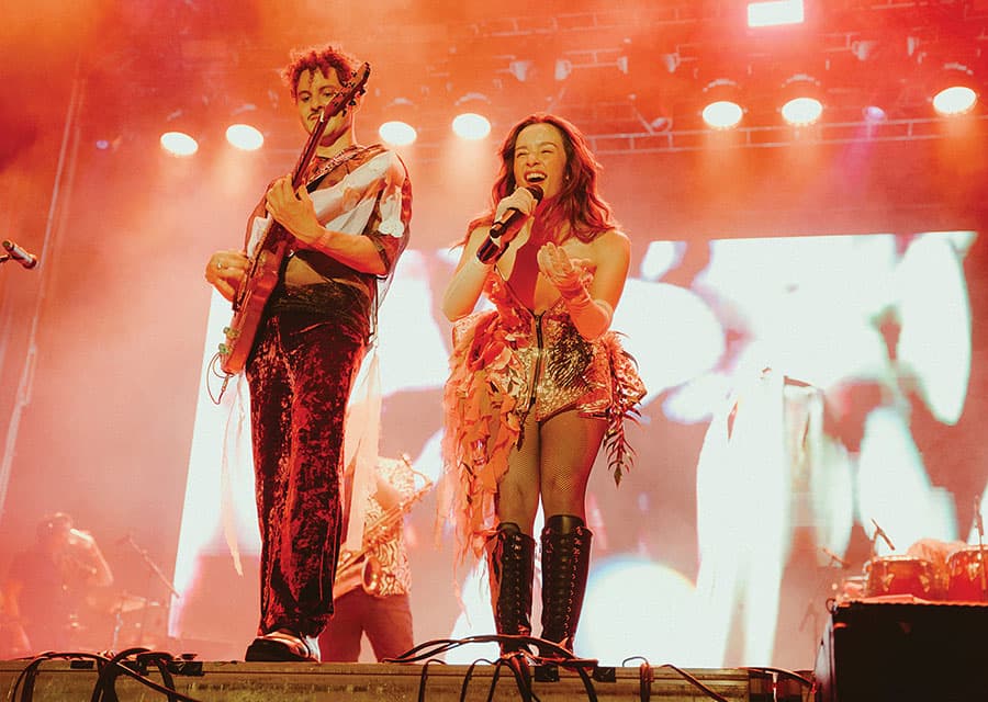 Duet on stage Man holding guitar, woman singing into a microphone with bright stage lighting behind them.