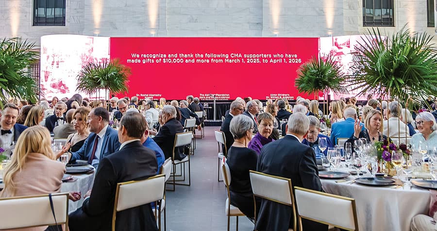 A large number of guests seated at tables in the museum atrium with a very large video display in the background. The text recognzes supporters of gifts of $10,000 and more on a bright red background. d
