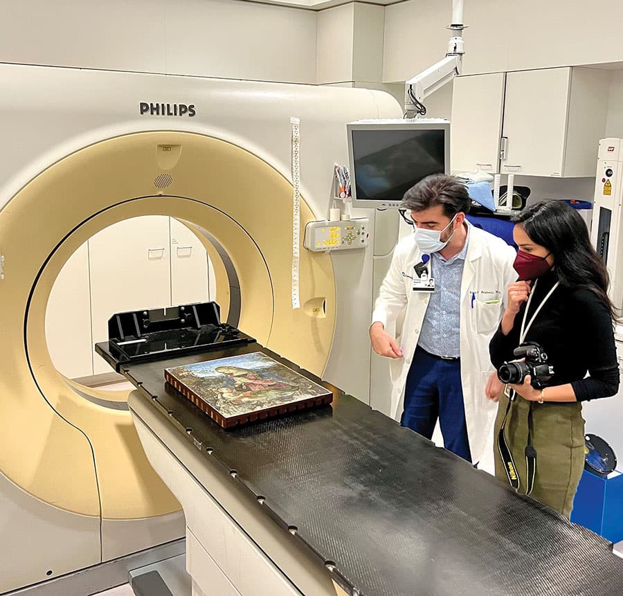 A painting on a platform prepared to slide into an MRI machine in a hospital. A man and a woman overseeing the activity, both wearing masks.