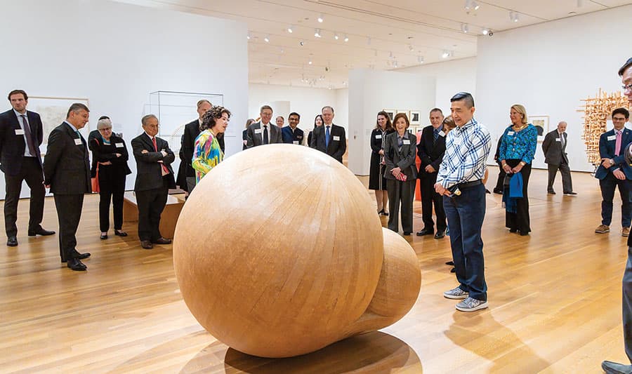 A large group tour gathered around a large sculpture, a sphere made of wood