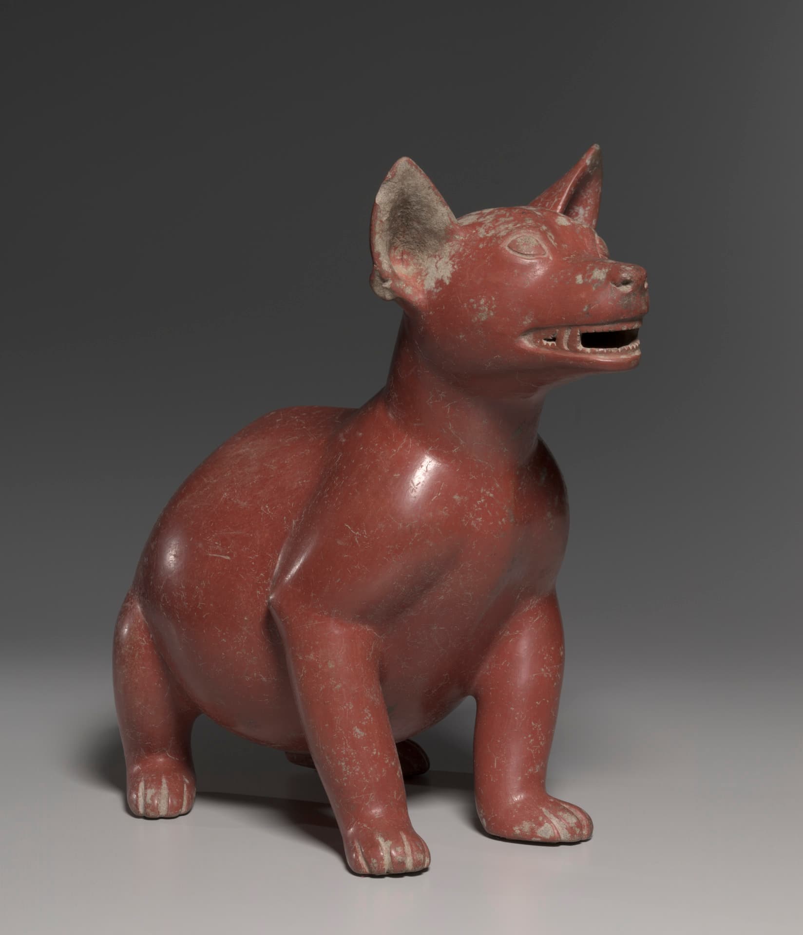 A red earthenware sculpture depicts a dog with a round body, crouching and looking up with its mouth open.