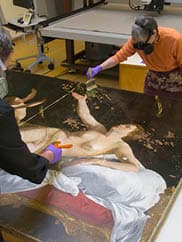 Two conservators work on a painting, laid out on a table between them.