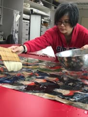 A conservator works on a painting, holding a bowl in one hand and brush in the other.