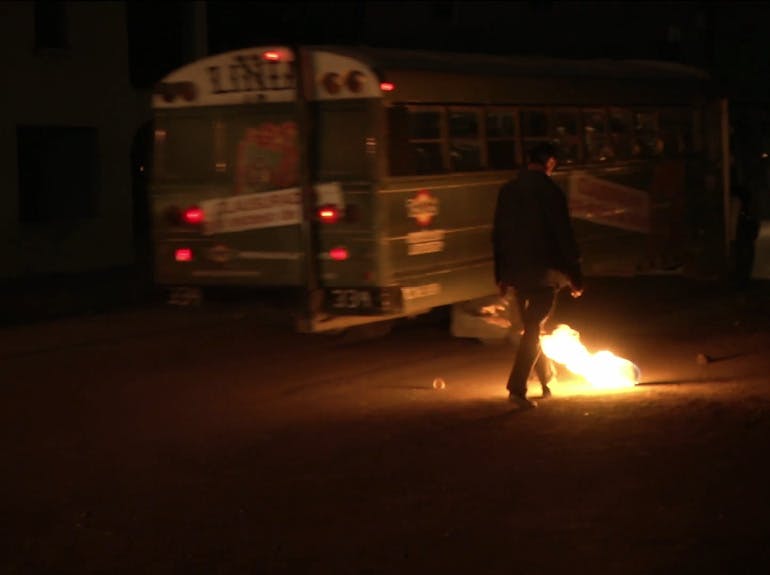 A video still of a person approaching a fiery object outside a bus at night