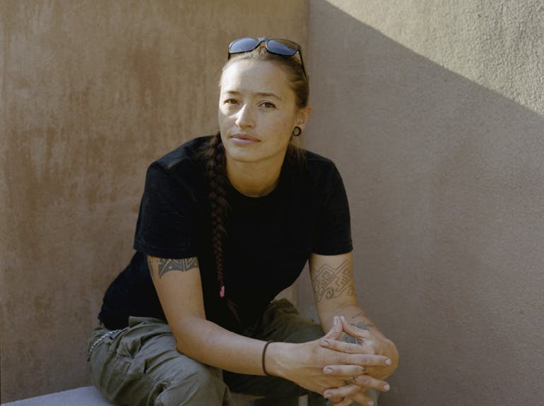 A photo of a seated woman in a black T-shirt with elbows on thighs and hands clasped