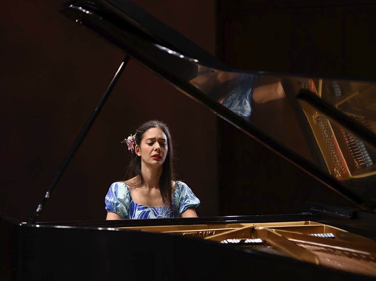A young woman with long dark hear and wearing a satiny blue dress emotively plays a grand piano.